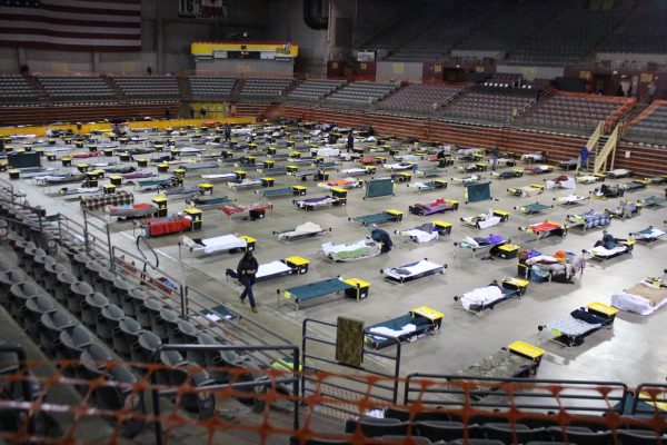 Cots are spread out on a stadium floor with empty bleachers spread around 