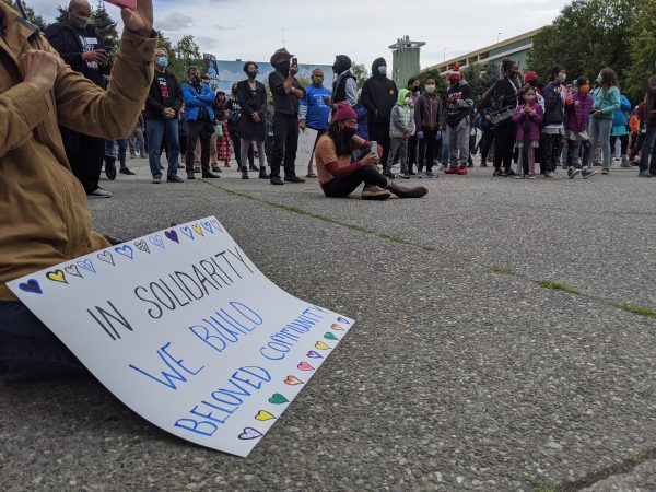 A rallygoer sits on the ground in front of a crowd of people with a sign that reads "In solidarity we build beloved community"