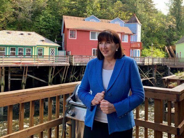 A woman in a blue suit coat holds her lapels. She hsas brown hair. She is standing on a deck with railings and wooden buildings in the background 