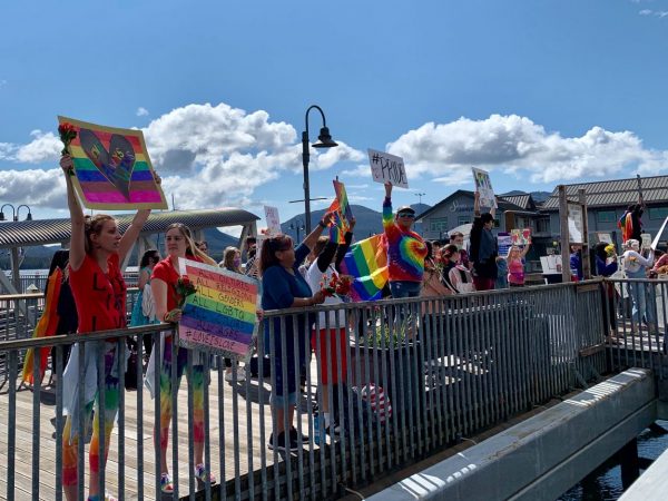 Supporters wave signs on a bridge on a sunny day. Many are wearing tie dyed shirts and rainbow colors. 