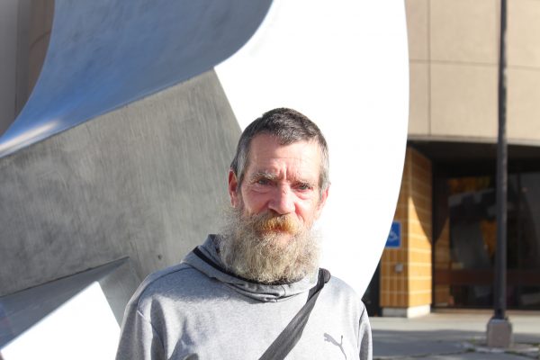 A white man in a grey hoodie and a greying beard stands in front of a stainless steel sculpture. 