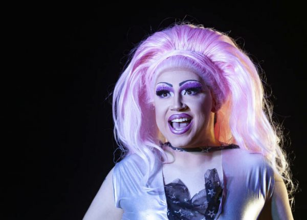 A man dressed in extravagant drag costume and pink wig sings on stage at a drag show