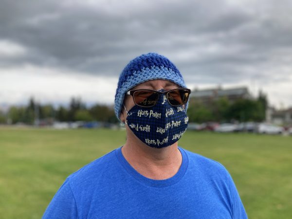 Portrait of a middle-aged white man man in a field wearing a blue beanie, sunglasses, and a mask with Harry Potter patterned across it