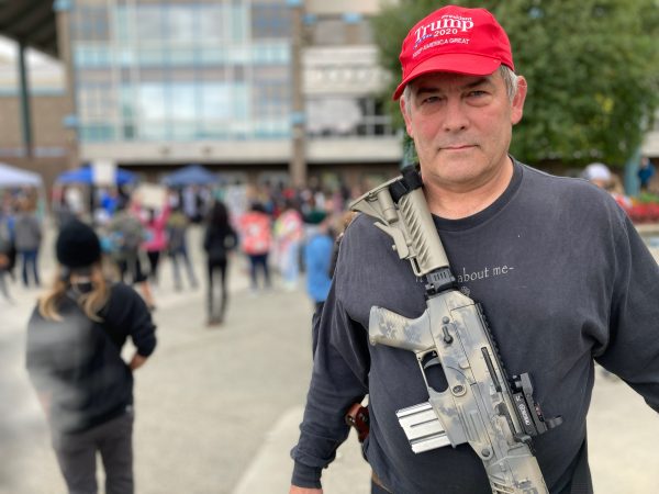 Portrait of a white older man wearing a red Trump hat with a rifle slung across his chest