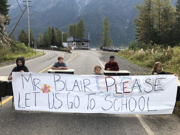 Schoolchildren sit ask a desk in the middle of the road behind a banner that reads "Ms Blair, please let us go to school". Mountains and trees are in the background. 