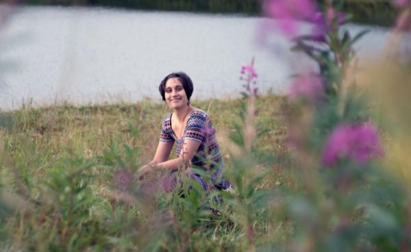A woman in a purple flowery dress sits cross legged in the grass behind fireweed and in front of a bay