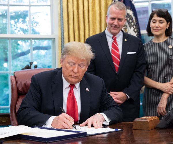 Trump sits at a desk, signing paper. A man and a woman stand behind him