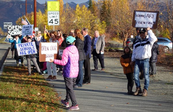 Protesters gather on the sidewalk in Anchorage with signs that read "Schools ARE Essential" and "Open our schools!"