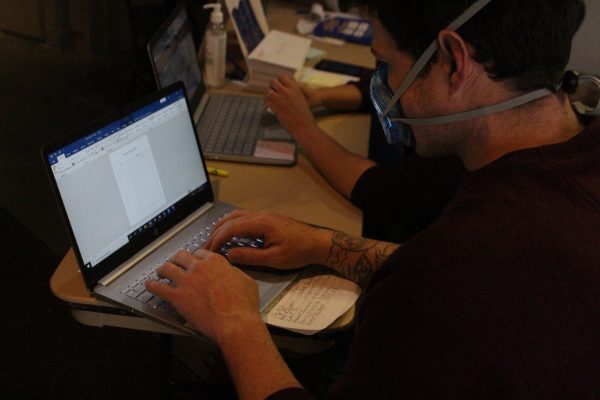 A white man in a blue mask and a red sweater tpes on a silver laptop. 