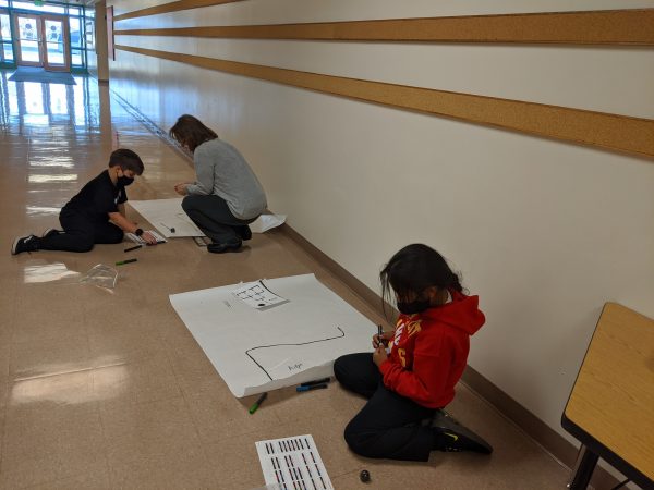 Two students and a teacher code small robots by marking colors on a piece of paper in a hallway in an elementary school