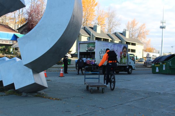 A man in an orange sweatshirt pulls a cart while riding his bicycled. Workers unload a truck in the background. 