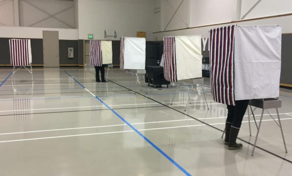 Voting booths spaced 6 feet apart in an open gym. People are voting inside them with their legs visible