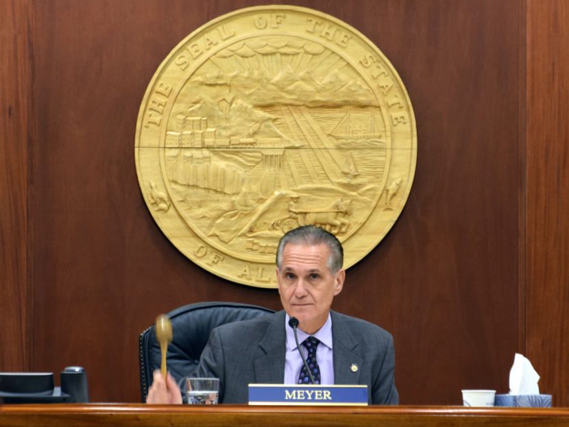 A white man in a gray suit beneath a gold state of alaska seal