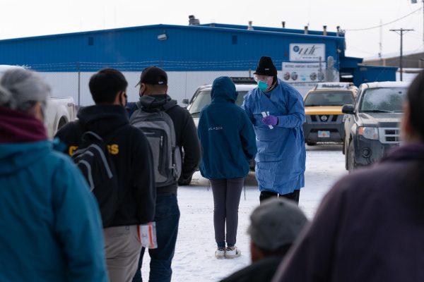 A health worker in a blue gown swabs travelers waiting in line outside the bethel airport
