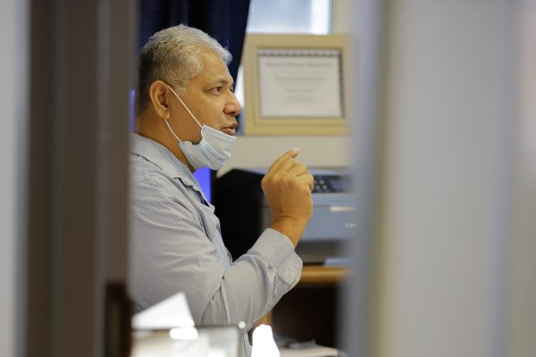 A grey-haired samoan man in a stripped button up shirt and a mask below his nose speaks in his office as seen through a crakc in the door