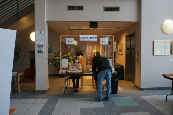 A man leans over his ballot at a table, separated from poll workers by a plexiglass panel.
