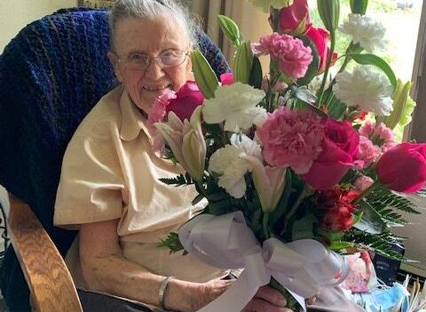 An elderly white woman sits in a chair, smiling, holding a bouquet of flowers