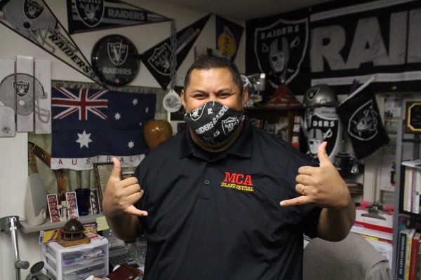A samoan man in a black collared t-shirt and a black oakland raiders mask gives a 'shaka' sign at his desk.