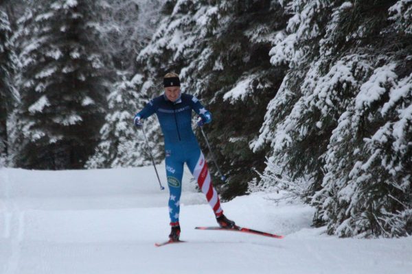 A skier in a blue and red white striped race uniform skis on a snowy trail with spruce trees around