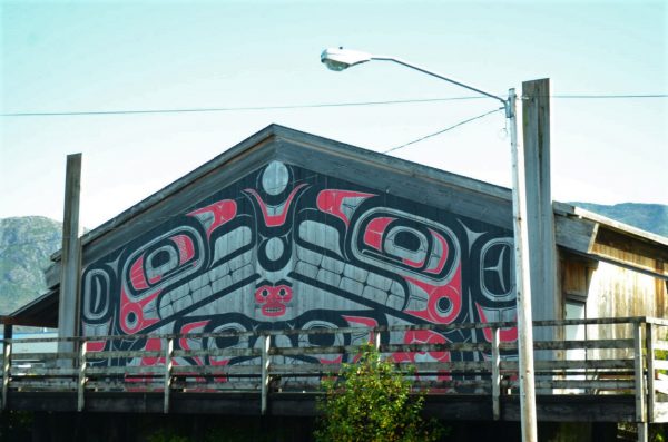 A structure, with mountains in the background, painted with Tlingit art.