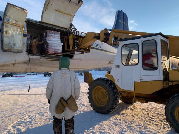In Tanana, a man in a parka waits to help a pilot unloads boxes of donated dog food from a small plane.