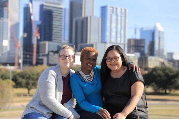 From left to right, a white woman, a black woman and an Alaska Native woman with their arms on each other's shoulders, smiling and posing for a photo with skyscrapers in the background.