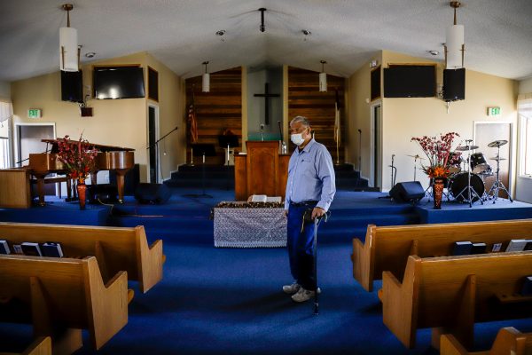 A grey-haired samoan man with a cane stands in the middle of several pews on a blue floor