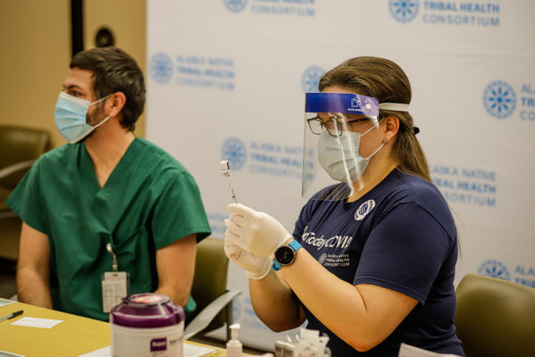 a person wearing a face shield and mask prepares to administer a shot to someone wearing scrubs and a mask