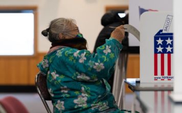 A woman in a flowery parka and a mamsk puts her ballot in a box