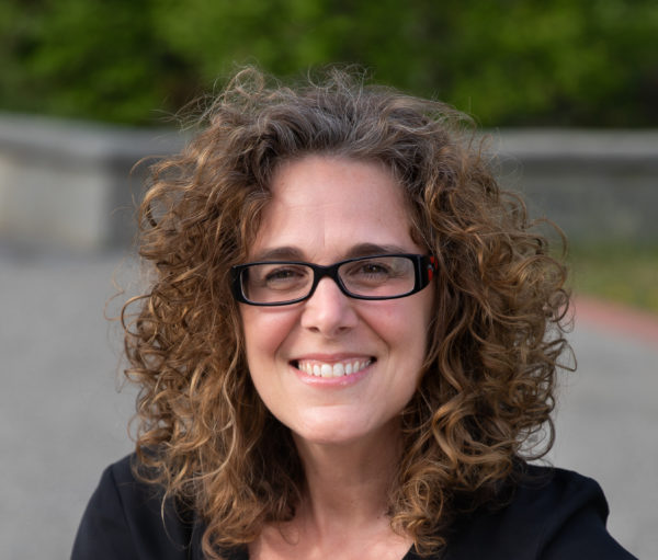 A headshot portrait of an white woman with curly brown hair wearing glasses