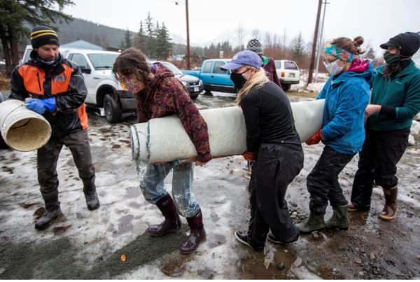 Several people carry a rolled up carpet in a muddy road