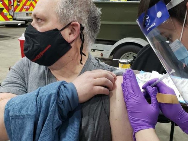 A man hols up his tshirt sleeve as he looks away with a mask and gets an injection