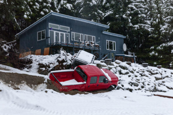 A truck below a house that is partially on its side and covered with light snow