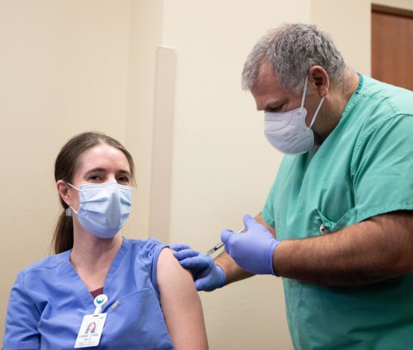 A woman wearing scrubs and a face mask receives a shot from a man in scrubs and a mask.