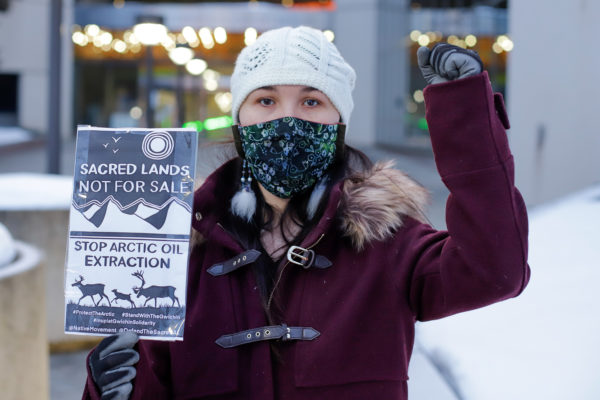 a person with their fist in the air holds a sign that says "sacred lands not for sale; stop arctic oil extraction"