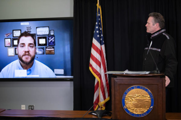 A white man at a podium looks at a video screen with another white man speaking