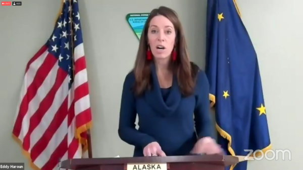 A white woman in a blue shirt speaks at a podium in fronnt of an american and Alaskan flag
