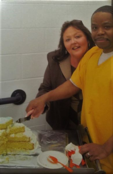 A,black man in a yellow suit holds the hand of a white woman as she cuts a cake in front of some white brick walls 