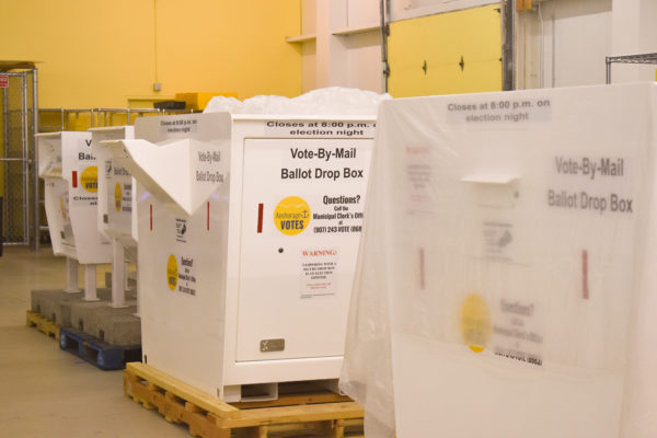 Large white boxes that read "Vote by Mail Ballot Drop Box" sit in a warehouse.