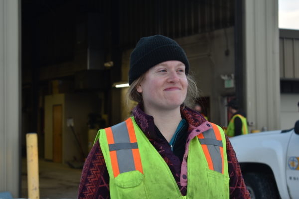 A woman wearing a plaid shirt, beanie and fluorescent work vest smiles in front of a warehouse.