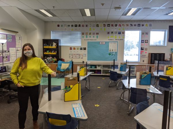 A woman stands in a classroom with her arm on a plastic barrier attached to a student's desk