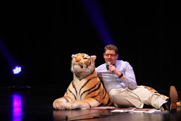 A white man sits on the stage floor with one arm around a stuffed tiger and the other holding a microphone