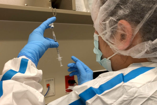 As seem from behind, a lab wowrker wearing ppe and gloves holds a syringe in their leeft hand