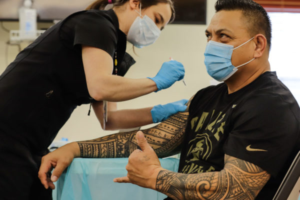 A man with tatooed arms gets a vaccine while giving a shaka symbol
