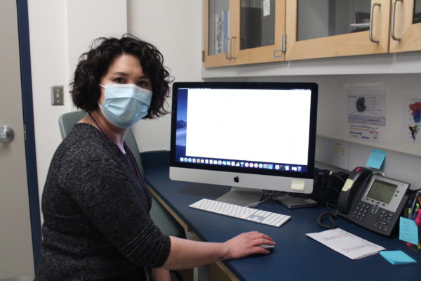 A woman with black, curly hair wearing a mask sitting at a Mac computer