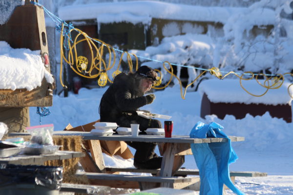 A man in a black snow outfit eats at a wooden picnic bench