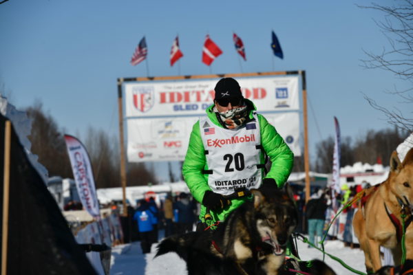 A musher in a lime green coat leaves the Iditarod starting line with his dog team.