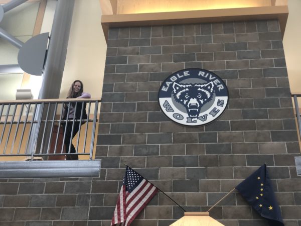 A girl stands at the top of a staircase next to a wall that has a crest on it and an American flag and Alaska state flag crossed beneath it
