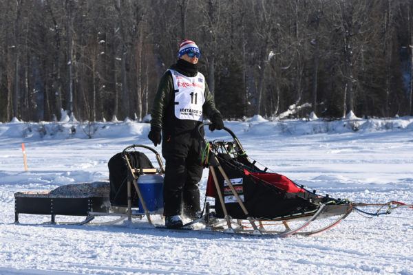 A musher on the back of a sled wearing a red-white-and blue hat.