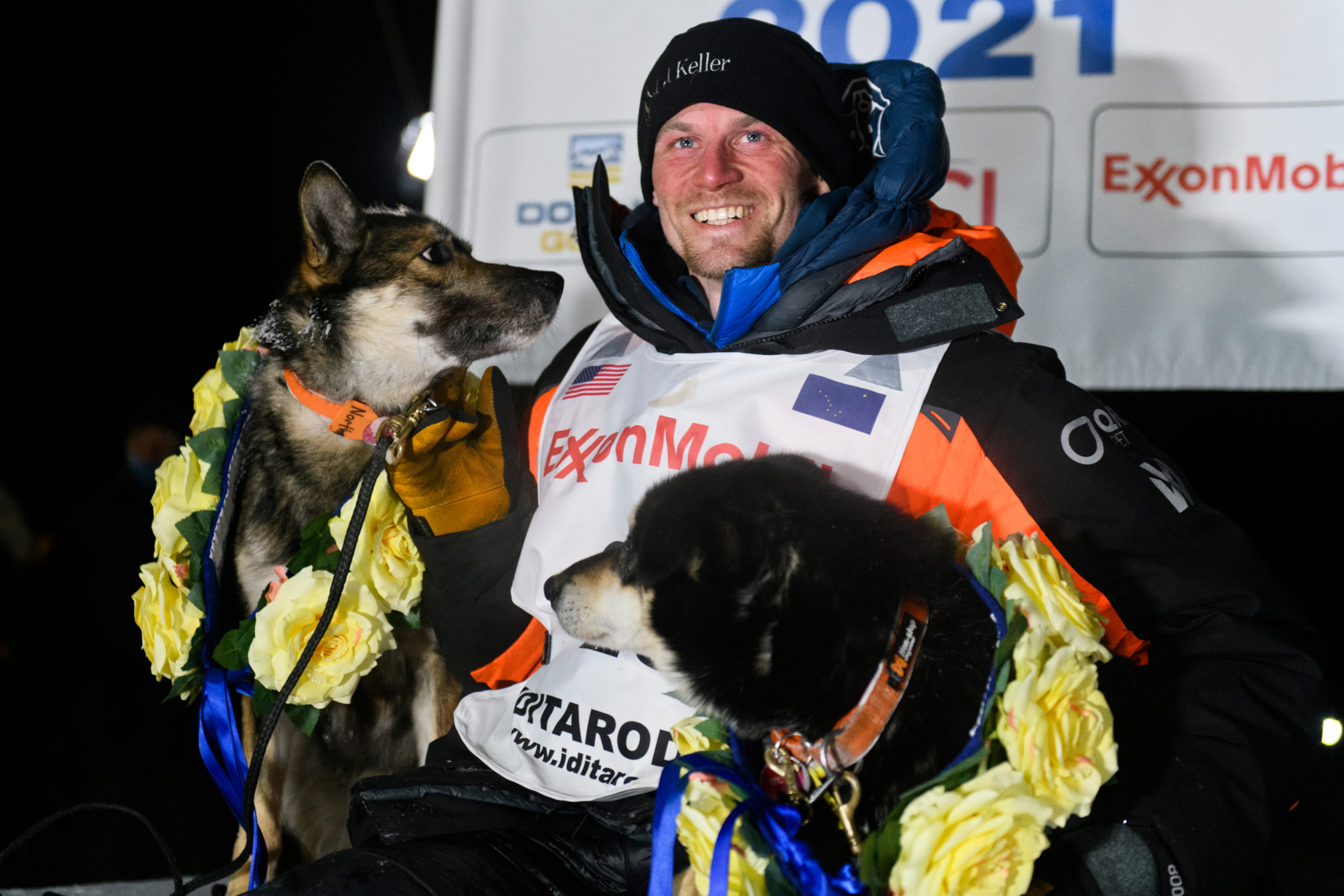 An Iditarod musher poses with two dogs wearing yellow flowers.